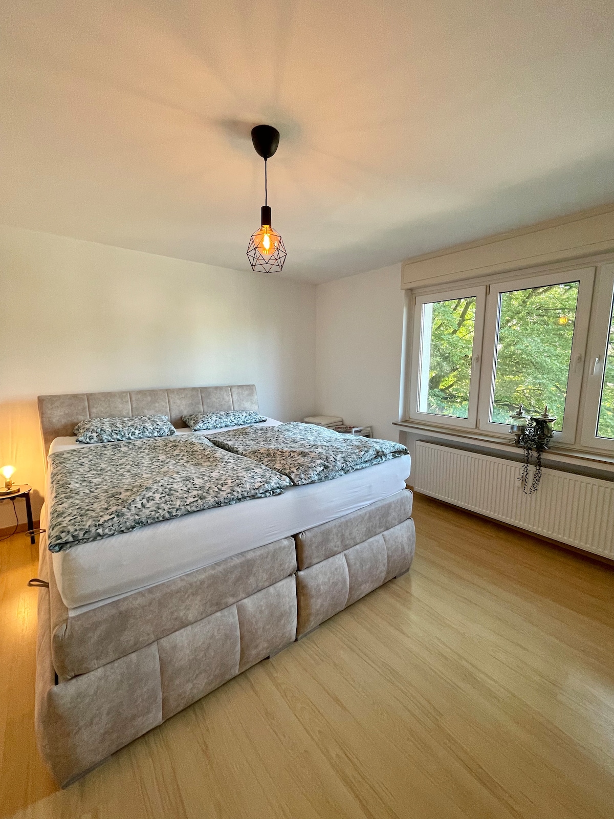 A bright bedroom is visible, featuring a spacious bed adorned with textured bedding. Natural light filters through large windows, framing views of greenery outside. A minimalist pendant light hangs overhead, contributing to the room's calming ambiance. A small nightstand is positioned beside the bed.