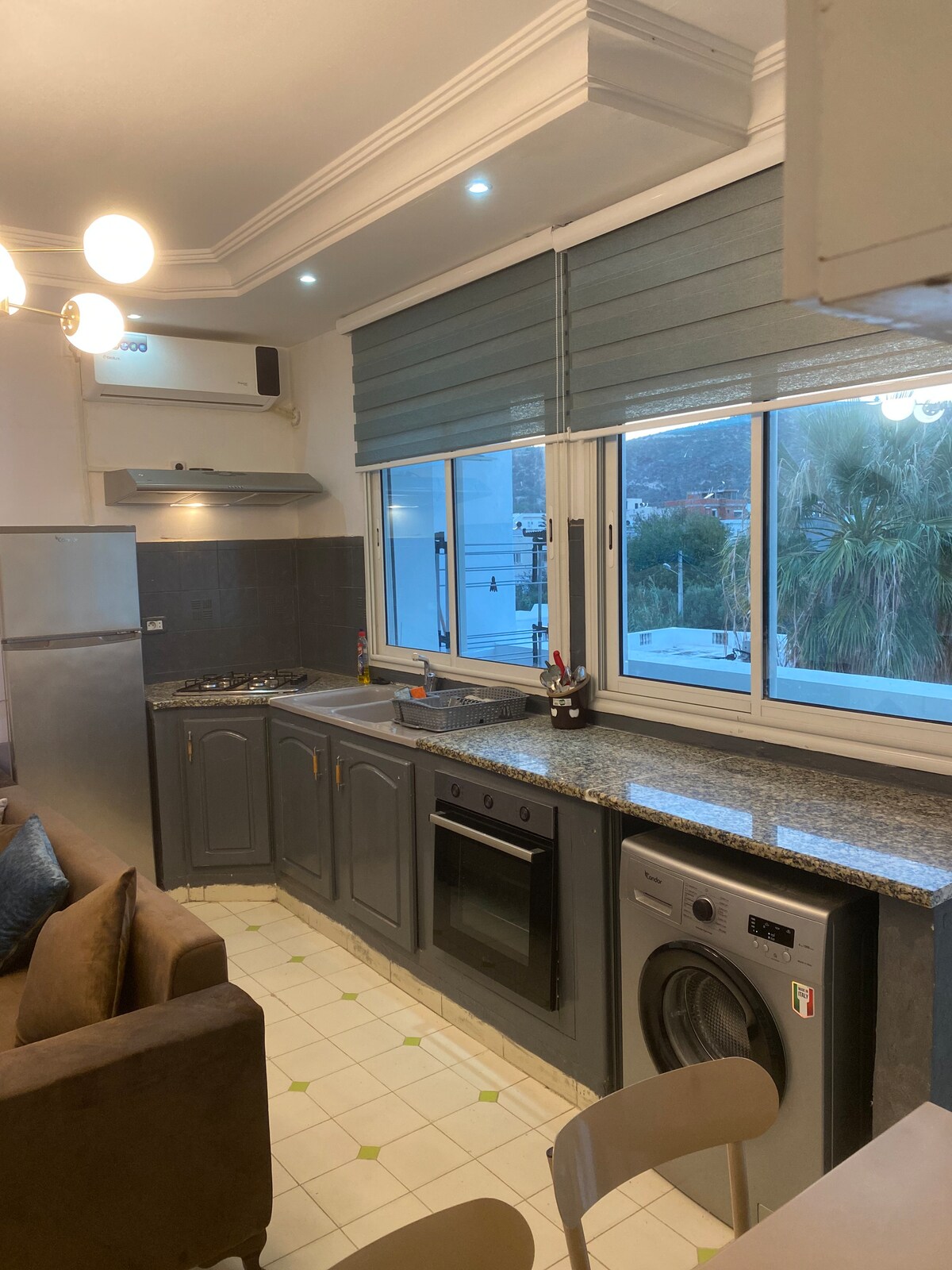 A modern kitchen features granite countertops and a comprehensive range of appliances, including a stove, oven, and washing machine. Large windows invite natural light, with views of the surrounding area, complemented by neutral cabinetry and light-colored flooring.