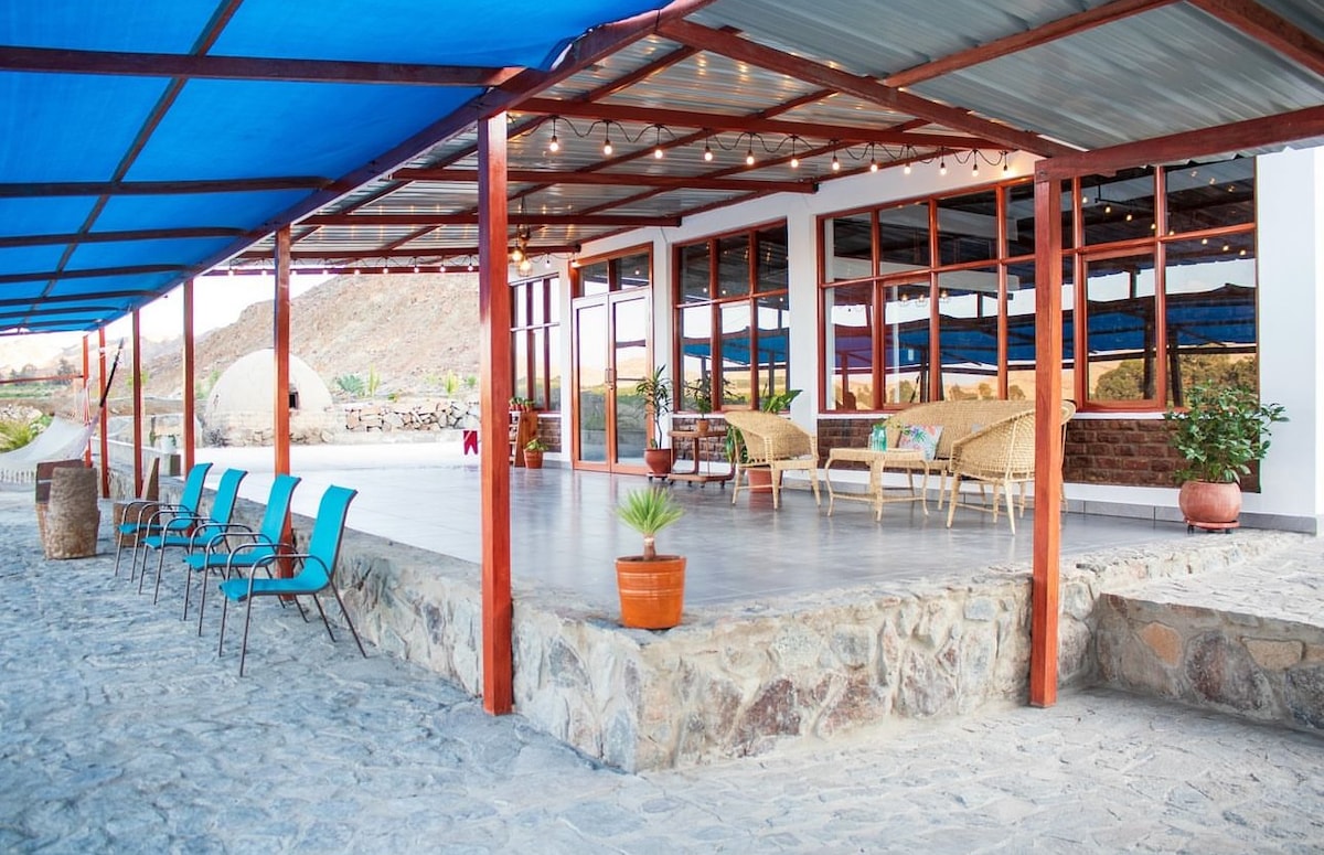 An outdoor seating area features a stone floor and a covered terrace supported by wooden beams. Comfortable seating arrangements are visible, along with potted plants. Blue fabric overhead provides shade, while large windows allow for natural light to fill the interior space.