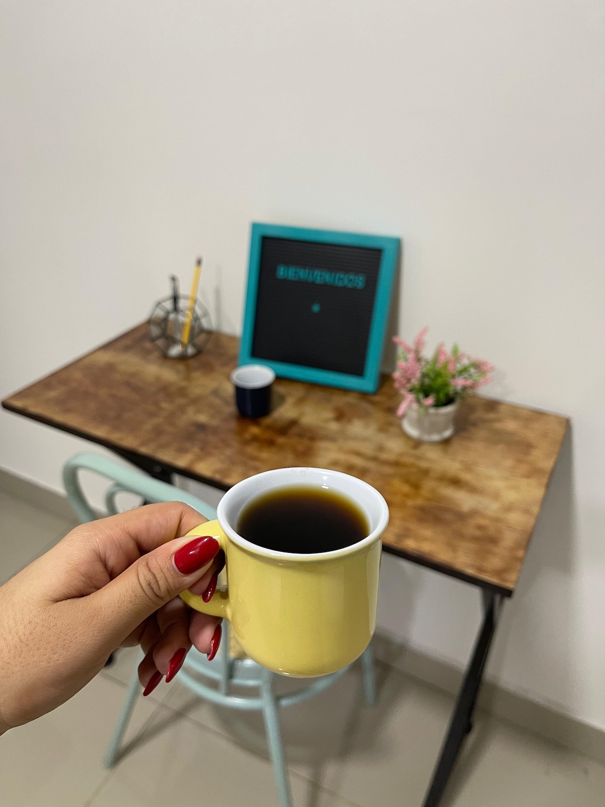 A small yellow cup of coffee is held in front of a wooden table. A welcome sign in a blue frame is positioned on the wall, accompanied by a small vase of flowers and a dark blue cup on the table. The overall setting conveys a simple and inviting workspace.