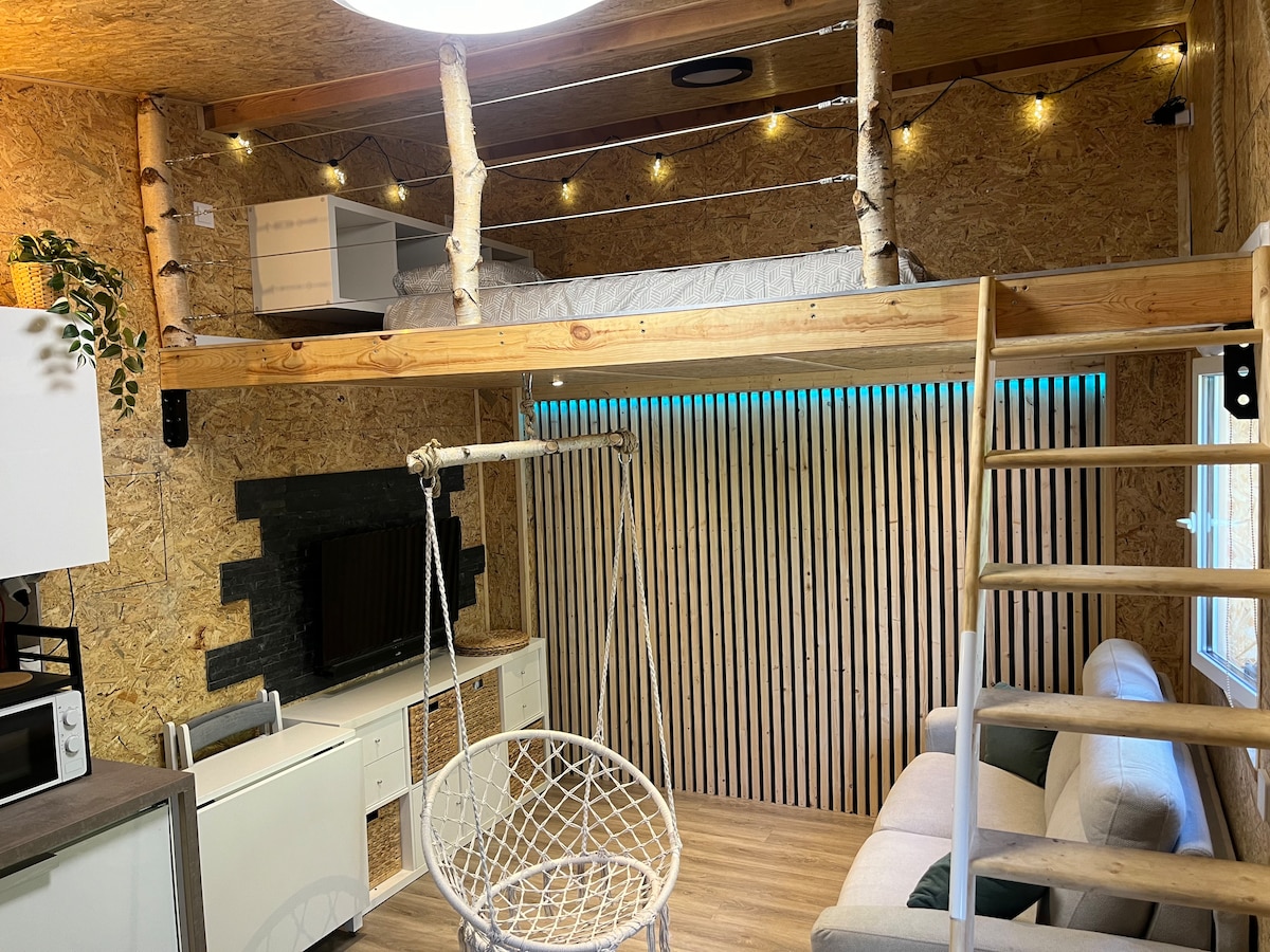 An open living space showcases a cozy seating area with a light-colored sofa and a hanging chair. A wooden loft is visible above, featuring a bed with soft bedding. Ambient lighting accentuates the natural wood walls, while a sleek kitchen area completes the scene.