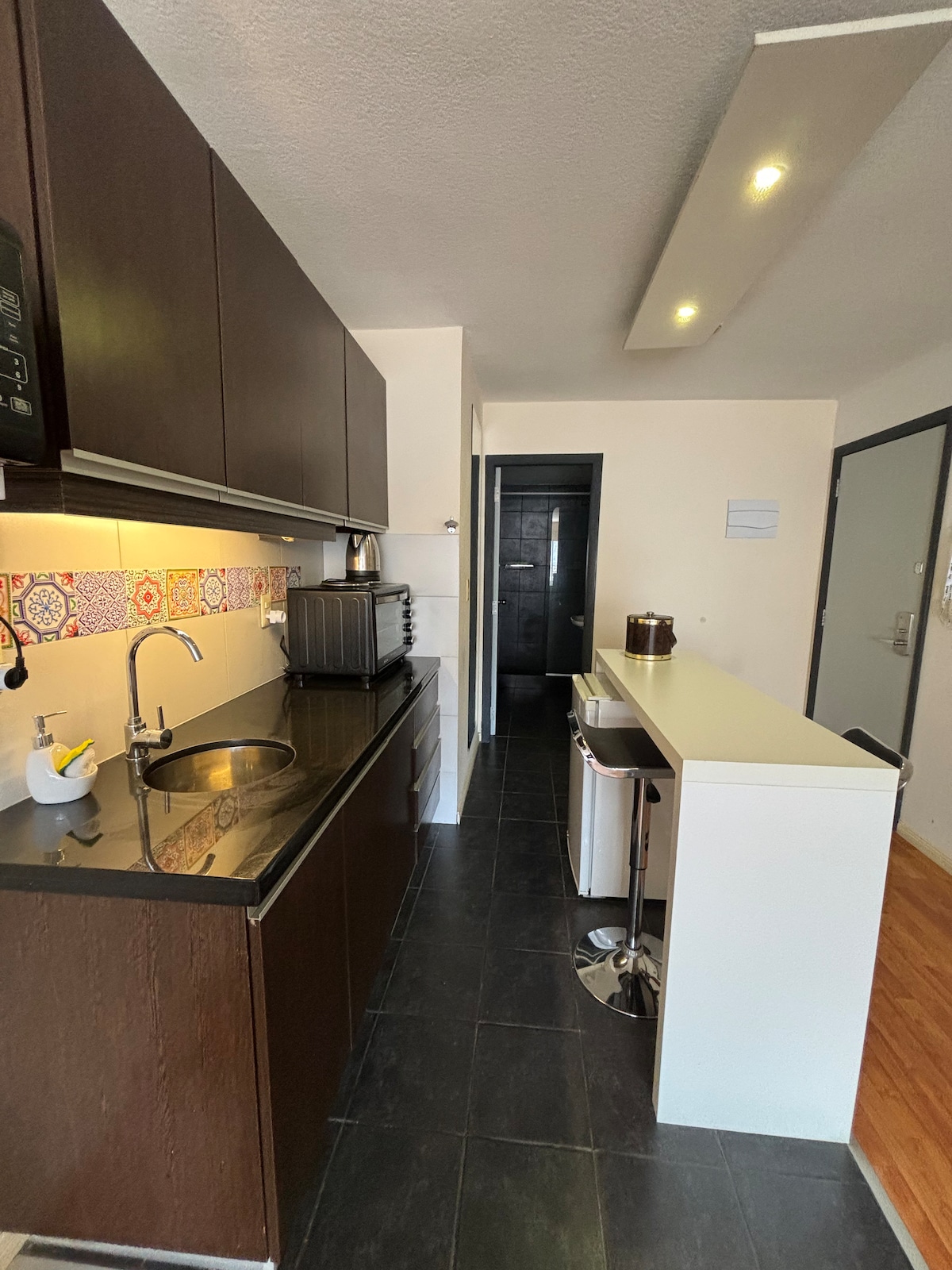 A modern kitchen area is shown with dark cabinetry and a black countertop. Kitchen appliances, including a kettle and toaster, are neatly arranged. A sleek bar counter with two stools provides a place for casual seating, while a door leads to the adjoining space.