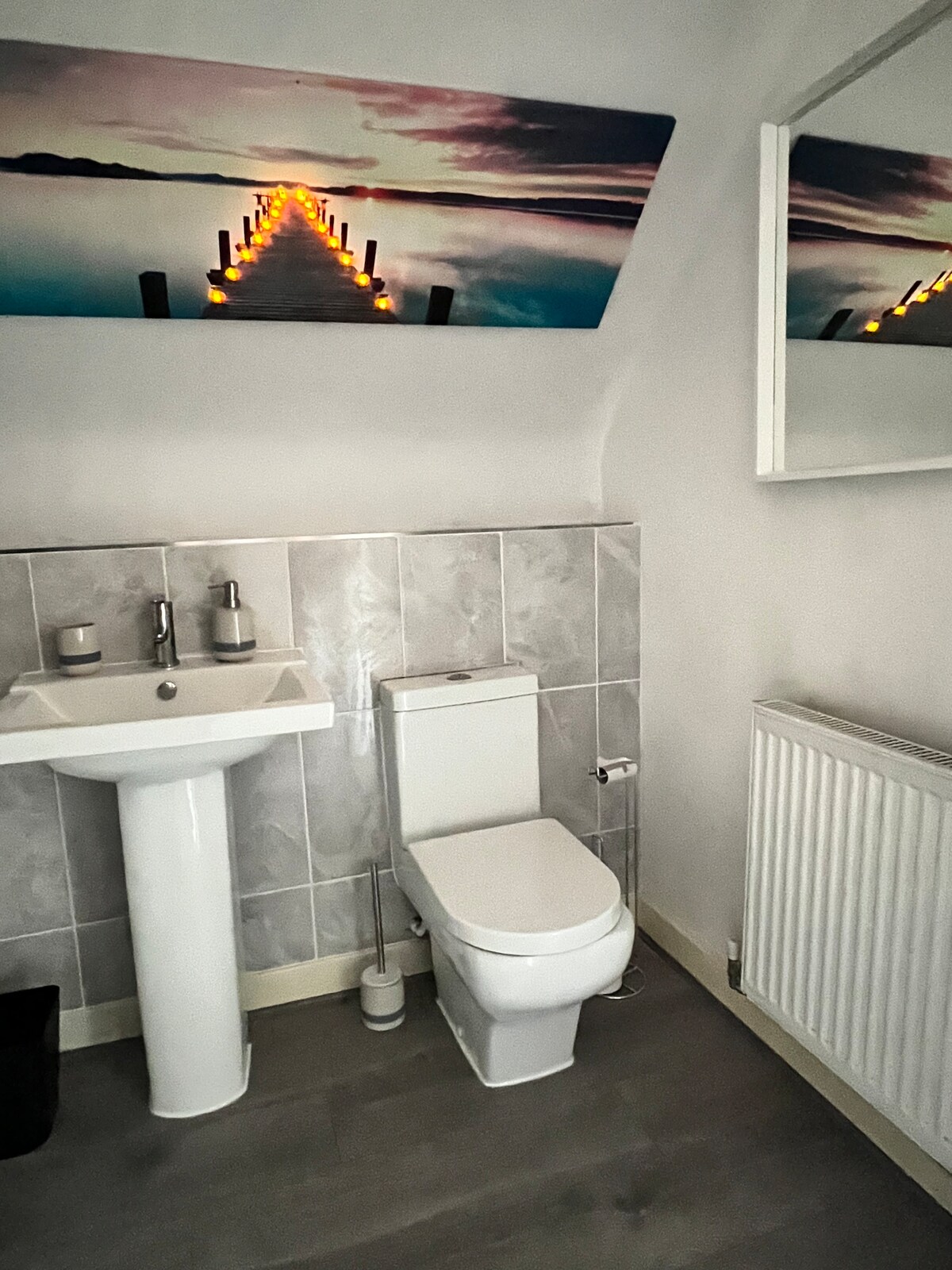 A contemporary bathroom is shown, featuring a white pedestal sink and a wall-mounted mirror above. A modern toilet is positioned next to the sink, with a tiled wall providing a clean appearance. A scenic image of a pier is displayed above, enhancing the space.