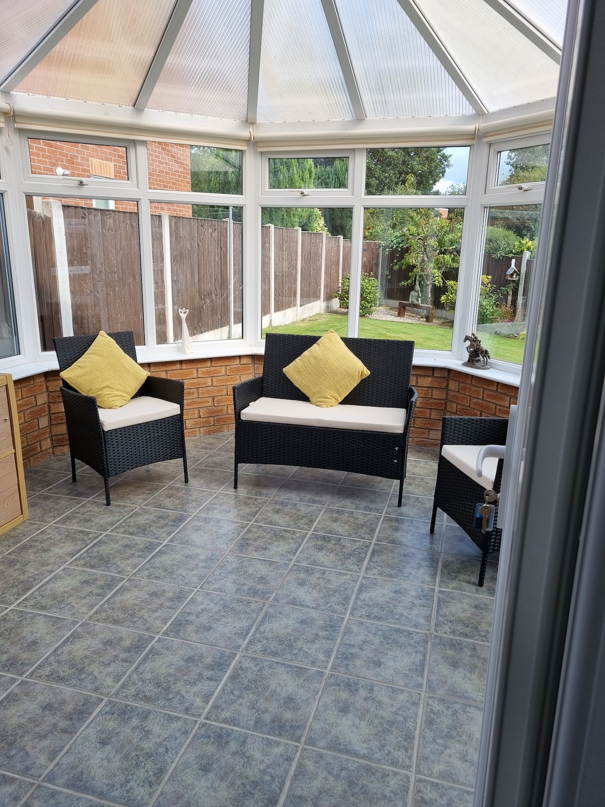 The conservatory features a tiled floor and large glass panels, providing ample natural light. Two wicker chairs with soft cushions and yellow accent pillows are positioned near the brick wall. A small storage unit is visible in the corner, and the garden can be seen through the windows.
