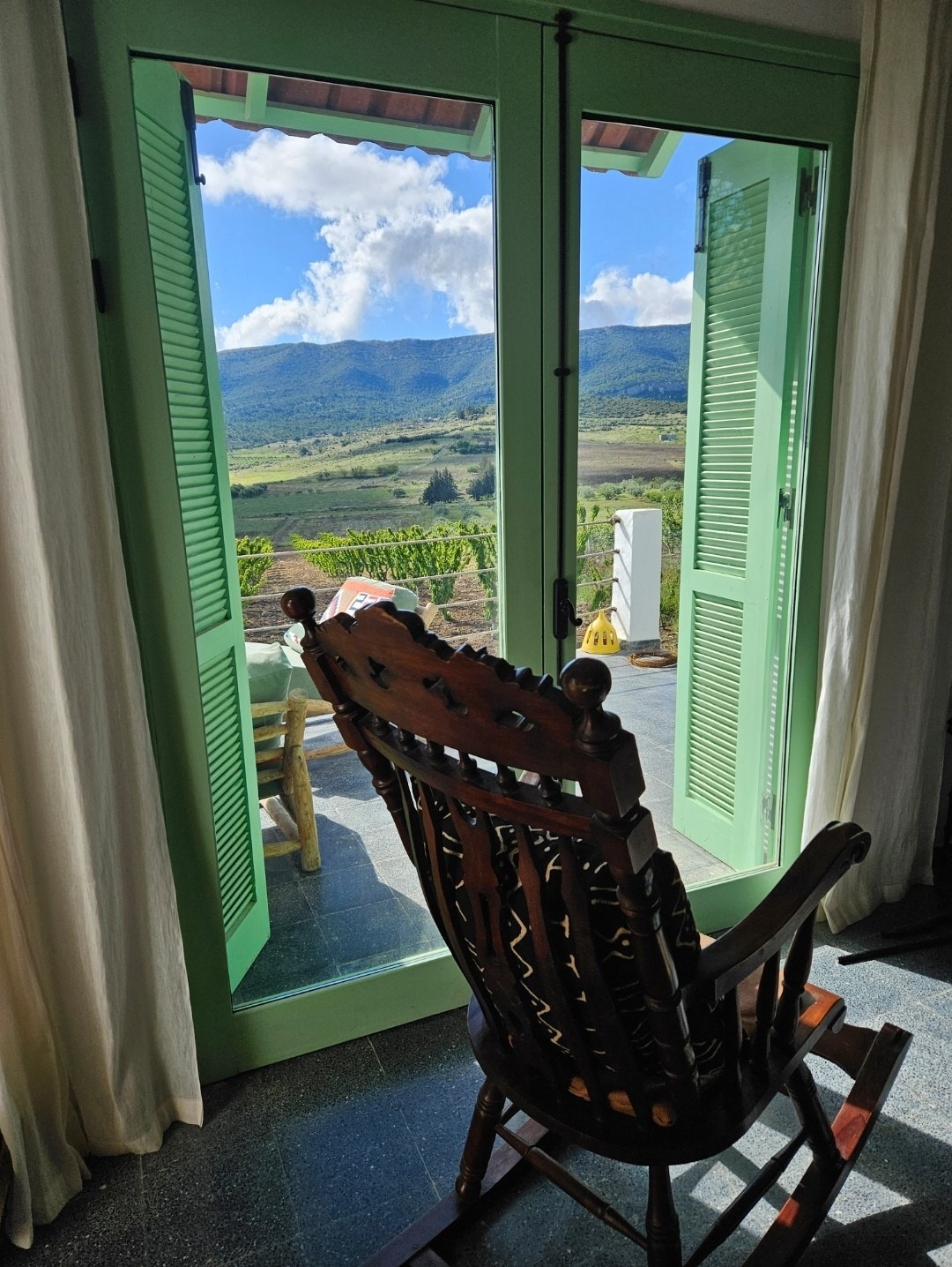 A rocking chair is positioned in front of open green shutters, framing a stunning view of the mountain landscape. Soft clouds contrast against the blue sky, while the patio area features additional seating, inviting moments of relaxation amidst nature.