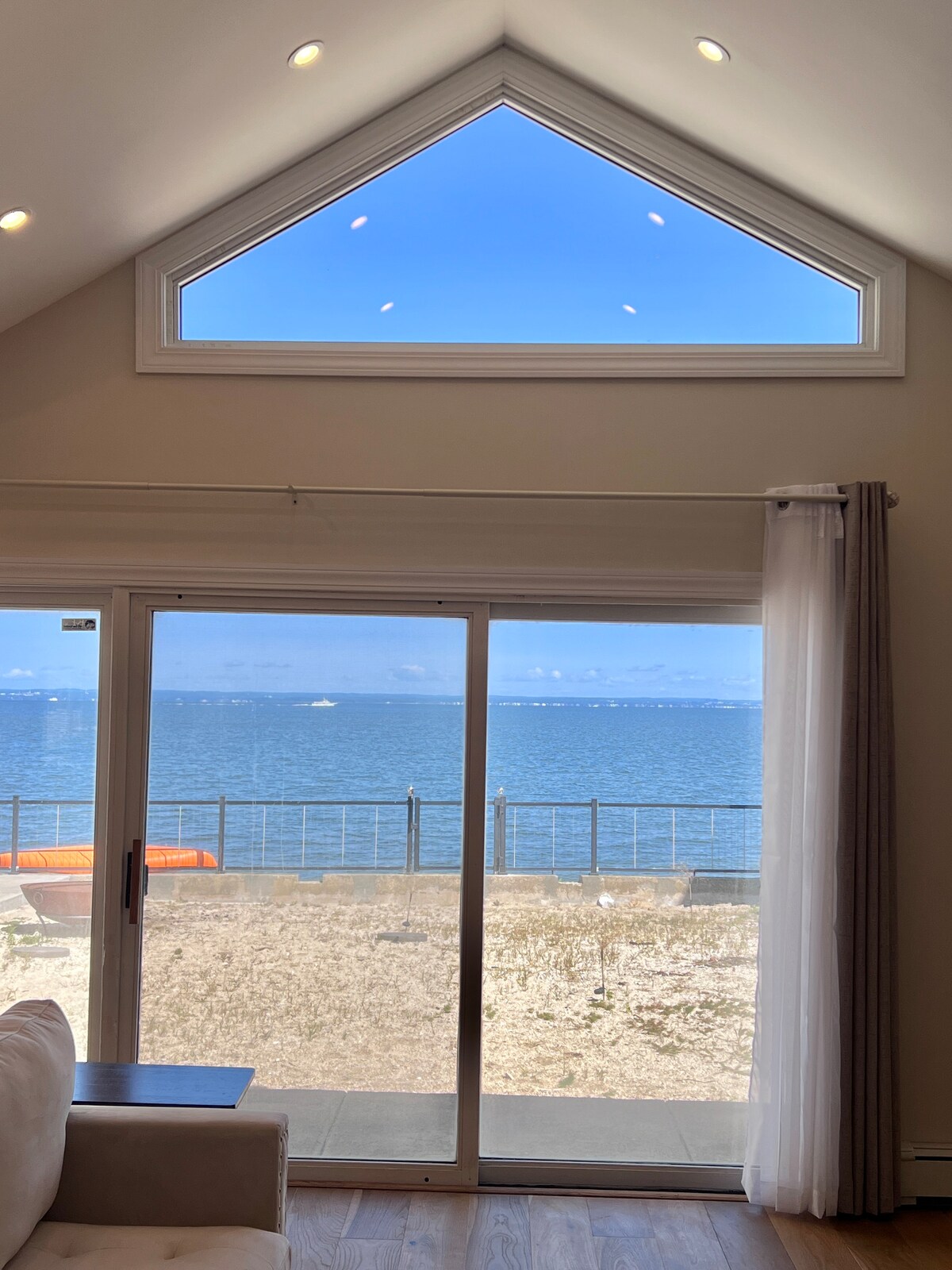 A large glass door offers views of the water, framed by light curtains. Natural light fills the room from the triangular window above, highlighting the modern interior. The peaceful waterfront can be seen through the door, enhancing the serene atmosphere.