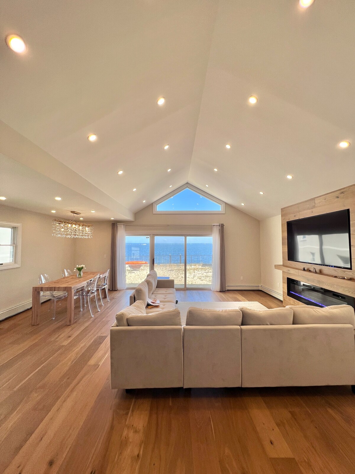 An open living area features a spacious layout with a light-colored sofa and a wooden dining table. Large windows offer views of the water, while recessed lighting brightens the room. A contemporary fireplace is positioned beneath a wall-mounted television, creating a cozy focal point.