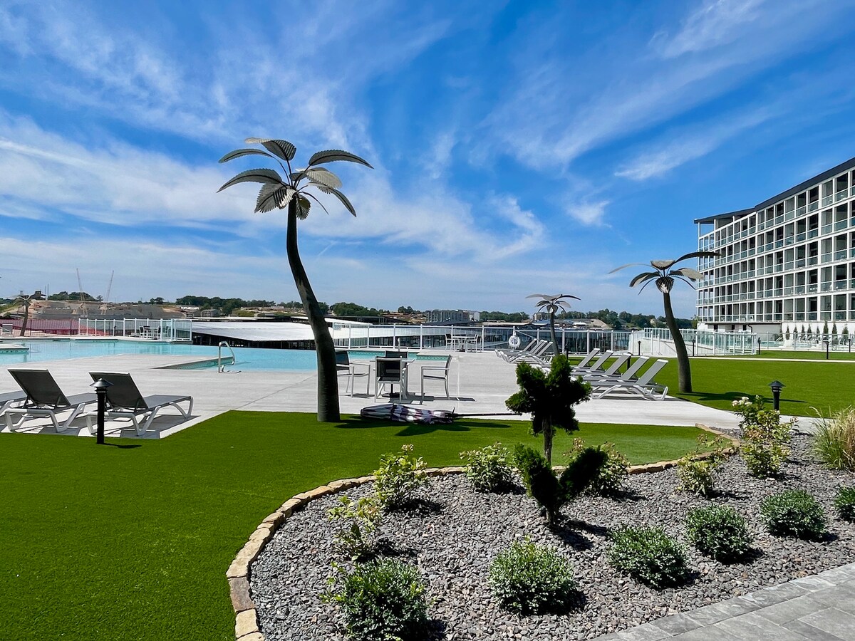 A vibrant pool area is showcased under a clear blue sky, featuring lounge chairs lined up near the water. Decorative palm trees add a tropical touch to the landscape, which is framed by neatly manicured grass and stone borders, enhancing the inviting ambiance.