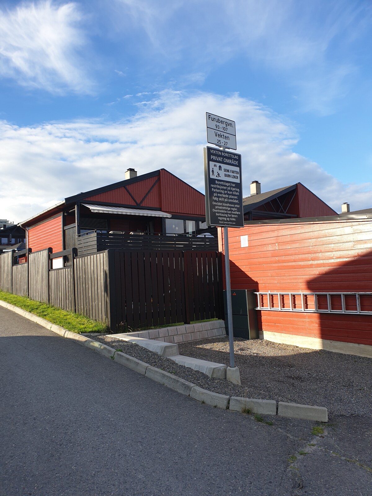 The image shows a peaceful neighborhood street featuring a sign indicating parking regulations. Alongside, red and black houses can be seen, separated by a wooden fence. The pavement is well-maintained, with a grassy area alongside the road.
