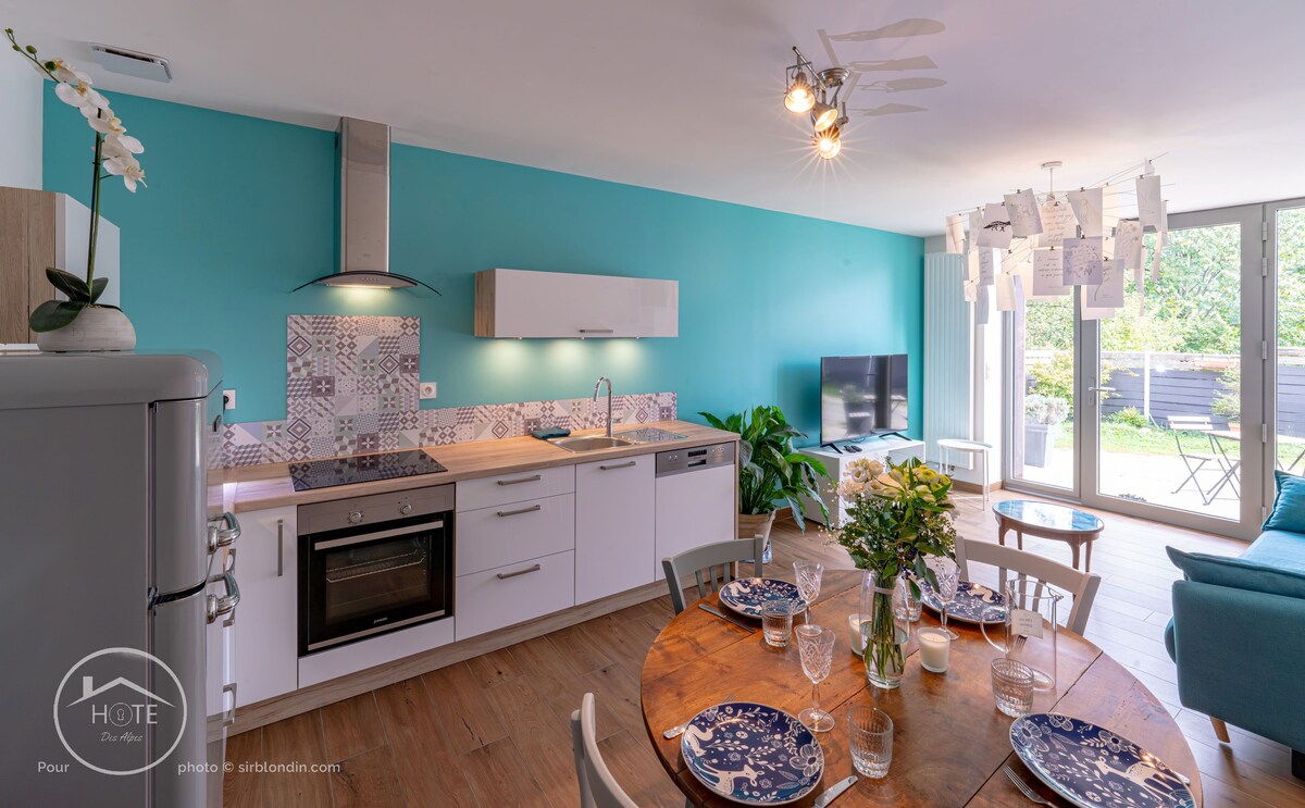 A modern kitchen area is seen alongside a dining space featuring a round wooden table set with plates, cutlery, and floral arrangements. The kitchen is equipped with white cabinetry and stainless steel appliances against an accent wall in teal. Large glass doors open to provide natural light.