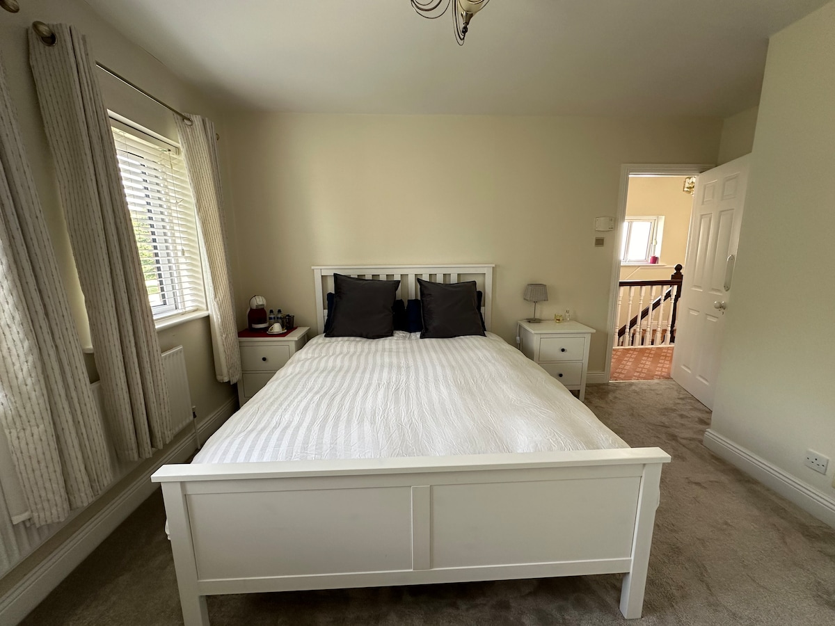 A bright sleeping area features a spacious white bed dressed in a textured blanket and flanked by two nightstands. Soft curtains frame a window, allowing natural light to illuminate the room. An open door reveals an entryway and access to the rest of the home.
