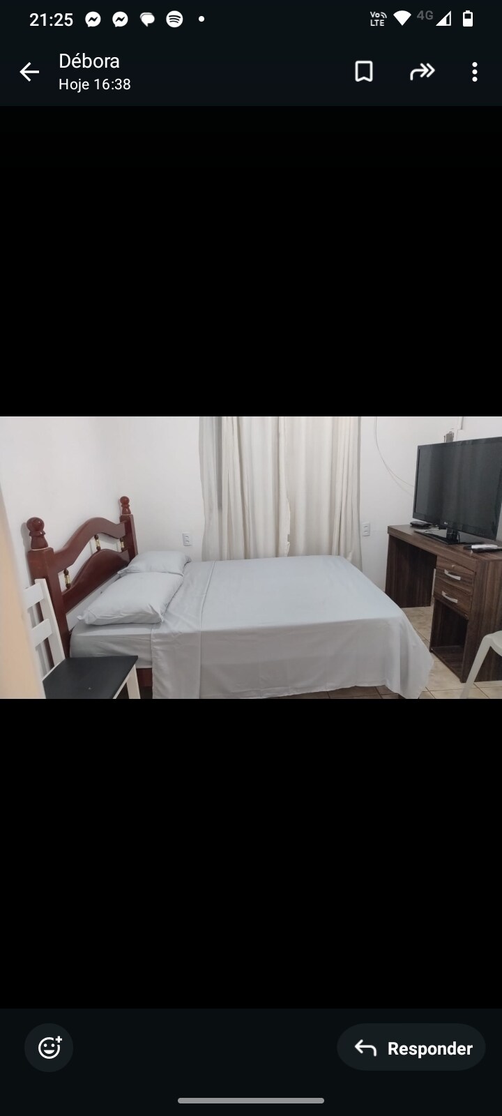 A simple bedroom is presented, featuring a wooden bed with a light-colored bedspread. A television is mounted on the wall opposite the bed. Natural light filters through white curtains, complementing the neutral-toned walls. A small wooden nightstand and a chair are positioned beside the bed.