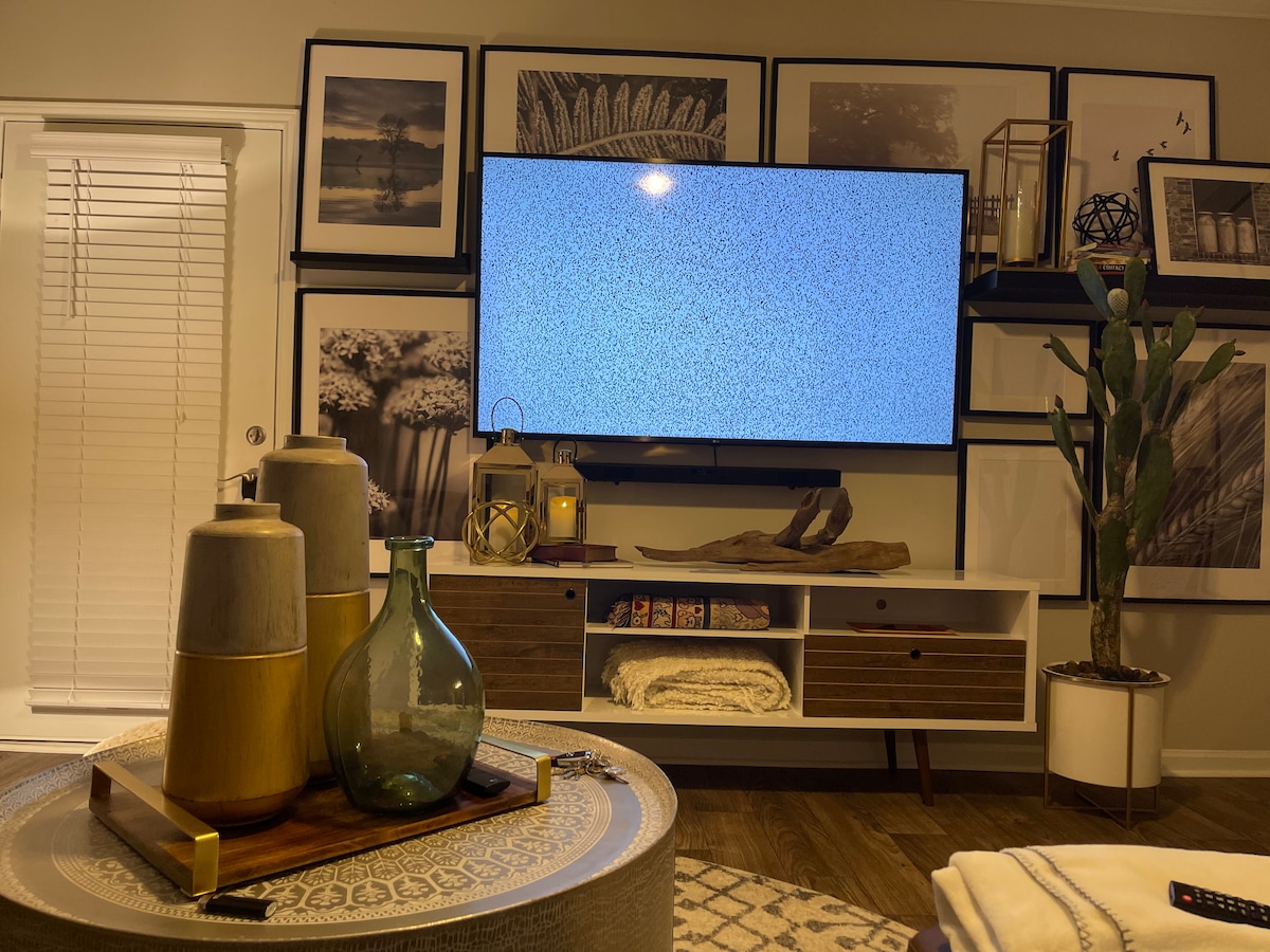 A modern living area features a large television atop a white media console, surrounded by a selection of framed artworks. A coffee table with textured surface holds decorative items, including glass vases and candles, beside a tall indoor plant, creating an inviting atmosphere.
