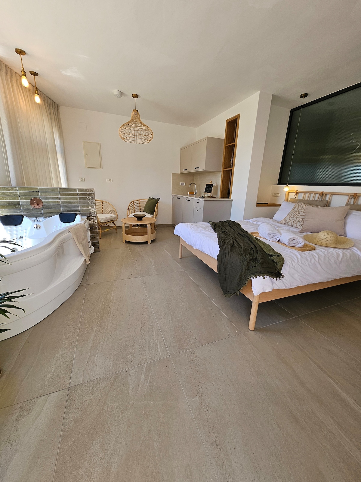 The room features a spacious bed covered with soft linens, accompanied by a cozy seating area with wicker chairs. A private spa tub is positioned nearby, and large windows let in natural light, creating a bright and inviting atmosphere.