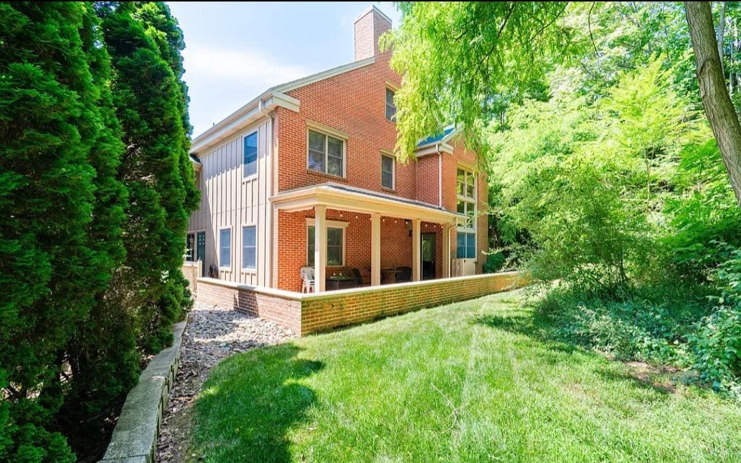 The exterior of the vacation home is shown, featuring a combination of brick and siding. Lush greenery surrounds the property, with a well-maintained lawn and shaded areas created by mature trees. The design includes multiple large windows, enhancing natural light.