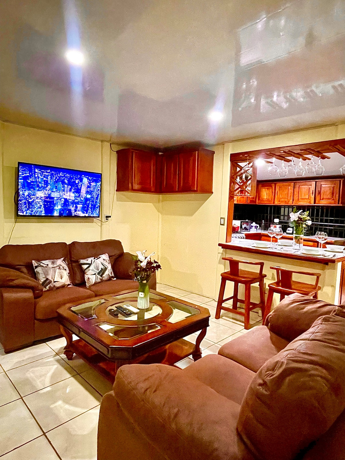 A cozy living area features comfortable brown sofas arranged around a glass coffee table. A television is mounted on the wall, providing entertainment. The kitchen is visible in the background, showcasing wooden cabinetry and bar stools. Soft lighting enhances the welcoming atmosphere.