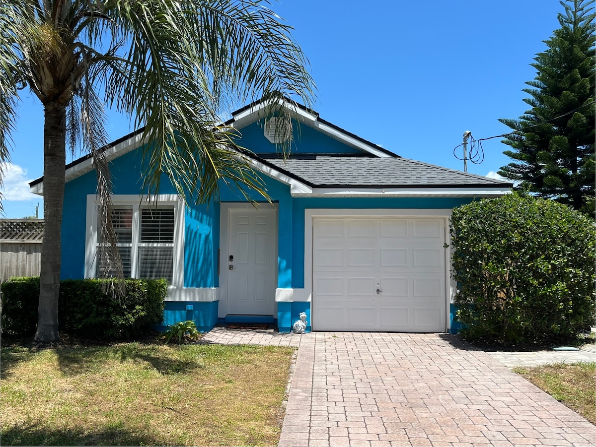 The exterior of a vibrant blue beach house is framed by lush greenery and palm trees. A well-maintained front yard features a brick pathway leading to a single-car garage. Clear blue skies complement the inviting character of this charming property.
