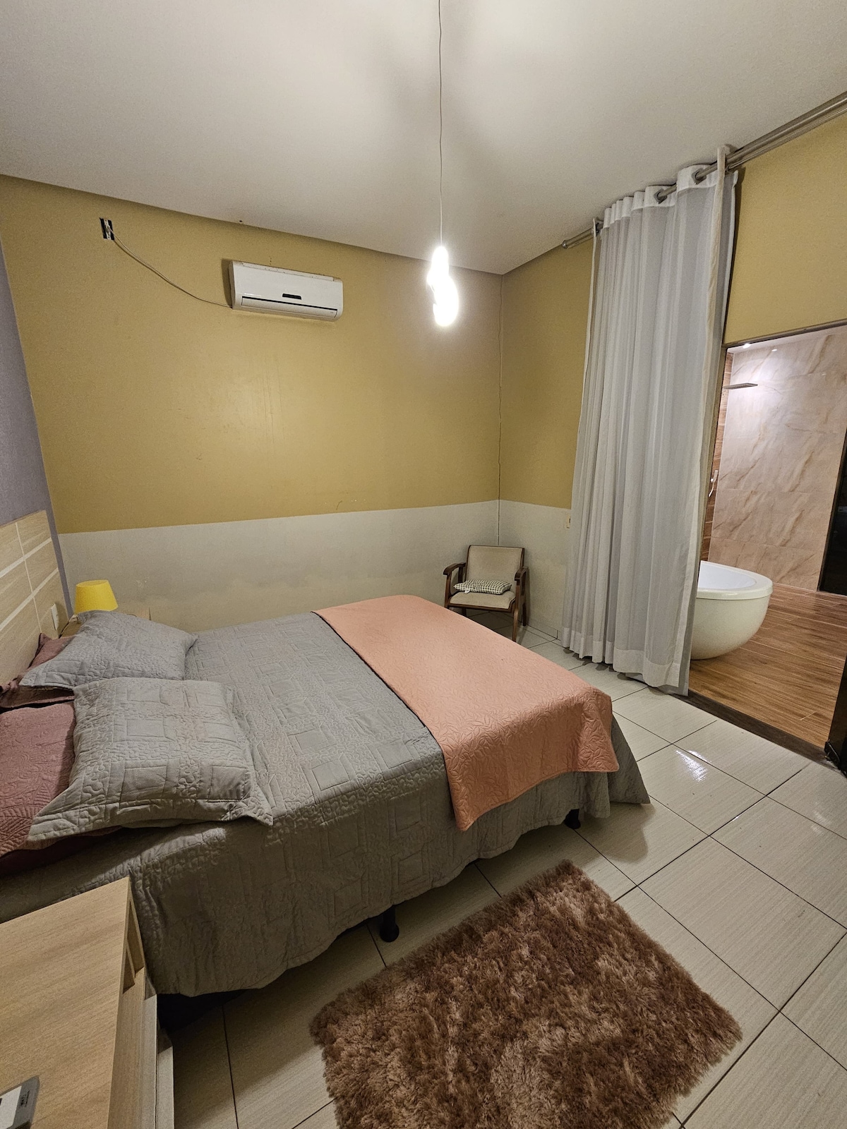 A cozy sleeping area features a queen-sized bed with a textured gray bedspread and a coral accent blanket. A small wooden chair is positioned beside the bed. Soft lighting is provided by a pendant lamp, and light-colored curtains frame the space. A modern bathtub is visible in the adjoining area.