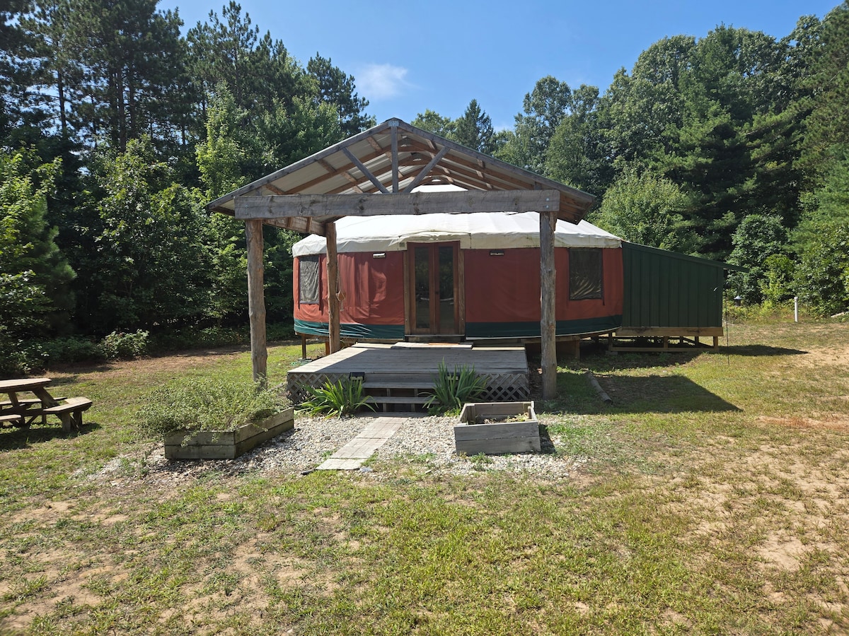 A yurt is situated under a canopy of trees, featuring a circular design with green trim. The entrance is framed by wooden posts and a small porch area, while picnic tables and garden beds are visible in the surrounding grassy area.