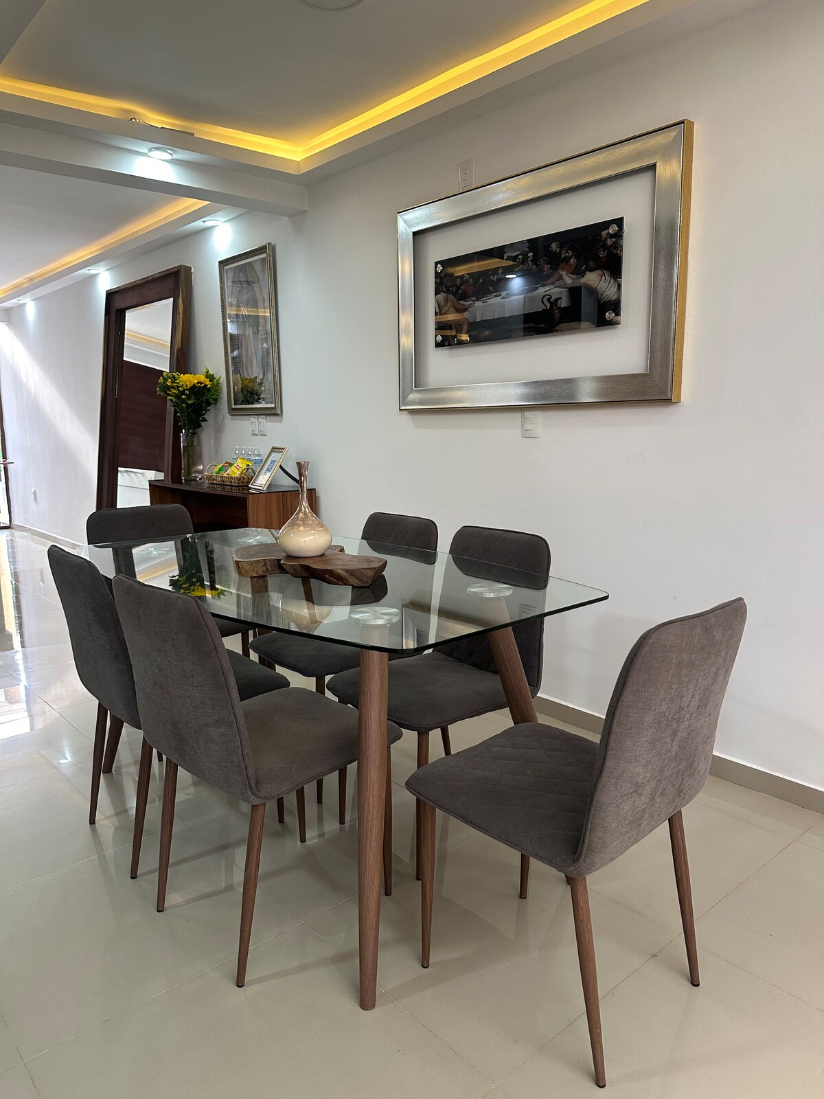 A dining area is depicted with a glass-top table surrounded by six upholstered chairs. A decorative bowl is placed on the table, while a piece of wall art is prominently displayed above. Soft lighting is provided by recessed fixtures along the ceiling.