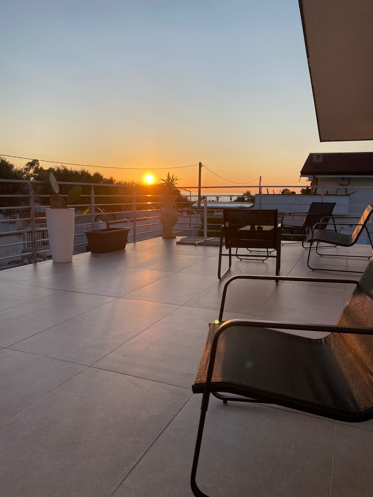 A spacious terrace is seen during sunset, featuring several chairs arranged for relaxation. Potted plants adorn the area, while the tranquil view of the horizon captures the fading light. The tiles reflect the golden hues of the sunset, enhancing the serene setting.