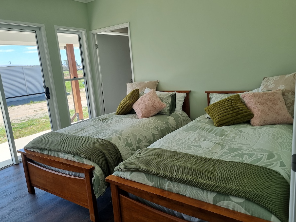 A bright and airy bedroom features two wooden-framed beds, each adorned with leafy patterned bedding. Decorative pillows in shades of pink and green add texture. Natural light floods the room through double doors, leading to an outdoor area with a view of the surrounding countryside.