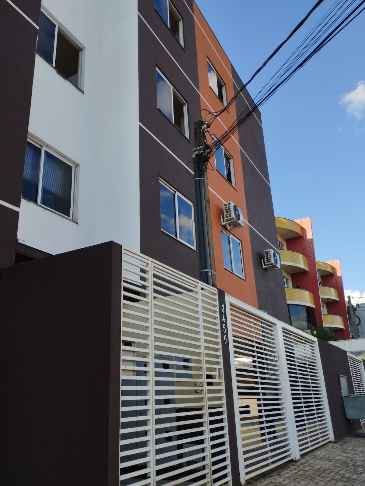 The building exterior features a combination of dark brown, white, and orange facades. Large windows are visible, allowing natural light to enter the interior. Air conditioning units are mounted on the outer wall. A gated entrance with vertical slats provides access to the property.
