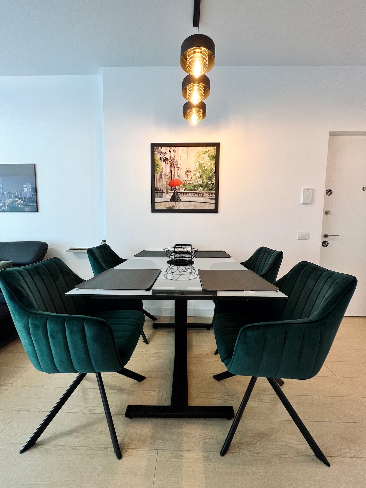 A dining area features a contemporary table surrounded by four green chairs. Above, three pendant lights provide illumination. A framed artwork depicting a city scene is mounted on the wall, adding a touch of character to the space.
