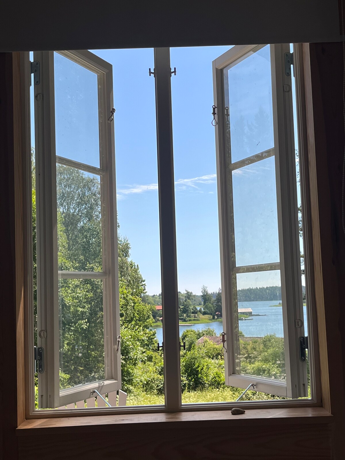 An open window with multiple panes provides a clear view of a serene landscape featuring greenery and a glimmering body of water. The bright blue sky can be seen through the window, enhancing the peaceful ambiance of the space.