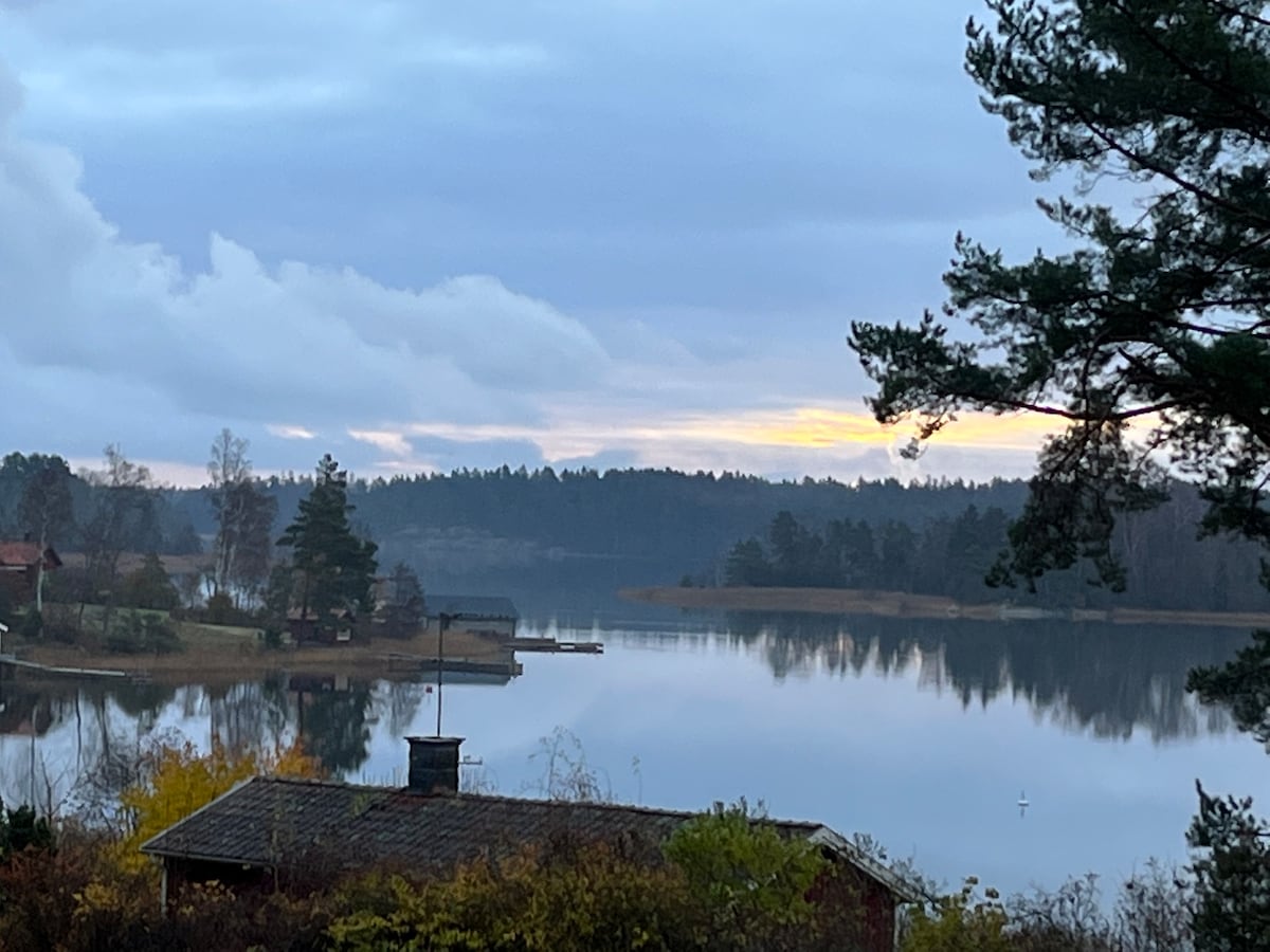 A serene view of a calm bay is captured, reflecting soft hues of the sunrise. Surrounding trees frame the water, while small structures dot the shoreline, enhancing the peaceful ambiance of the landscape.