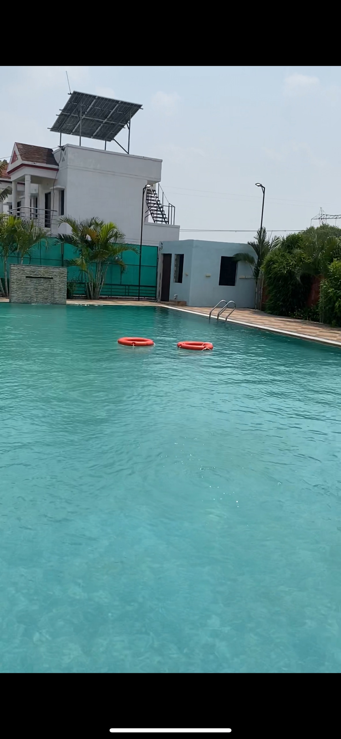 A large swimming pool is seen in a serene setting, featuring clear turquoise water. Two orange flotation devices float on the surface. Lush greenery lines the pool's perimeter, while a staircase and building are visible in the background.