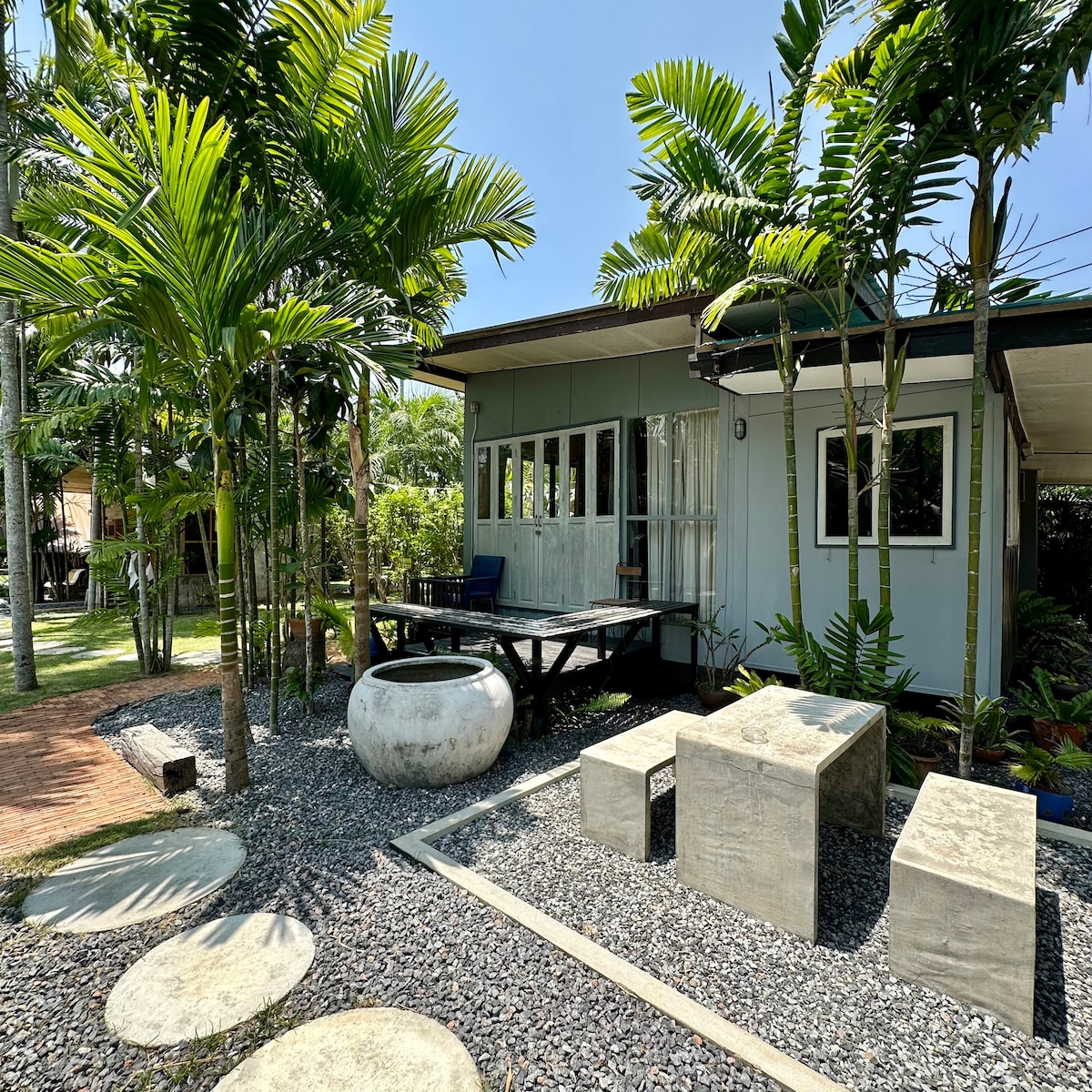 A modern dwelling is surrounded by lush tropical plants, including tall palms. A concrete seating area is visible, composed of a table and bench-style seats. The outdoor space features stepping stones leading through gravel, enhancing the serene garden atmosphere.