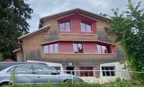 A three-story wooden house features a distinct red-trimmed exterior, with multiple large windows. The landscaping is natural, with greenery surrounding the base. A parked car is visible in front, with a driveway leading up to the entrance.
