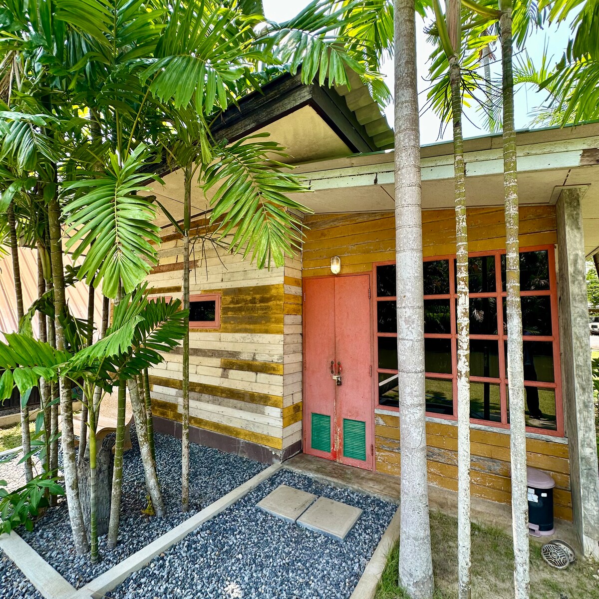 The exterior of a colorful wooden cabin is framed by tall palm trees. The structure features a vibrant red door and large windows, allowing natural light to fill the interior. A pebbled walkway leads to the entrance, enhancing the inviting atmosphere.