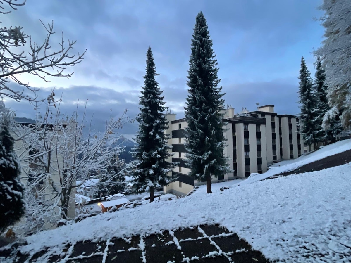 A winter scene captures a multi-story building set against a backdrop of snow-covered trees. The ground is blanketed in fresh snow, and the sky features soft gray hues, suggesting early morning light. The surrounding landscape showcases the beauty of the Alpine environment.