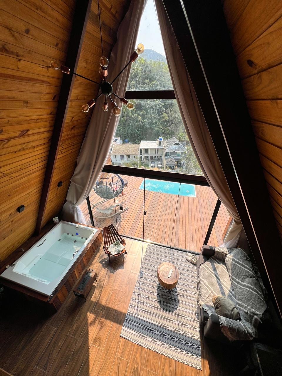 The interior features a cozy seating area with a modern hanging chair and a small coffee table. A large window offers a view of the outside deck and pool area, while a jacuzzi is positioned beside it, allowing for relaxation with natural light.