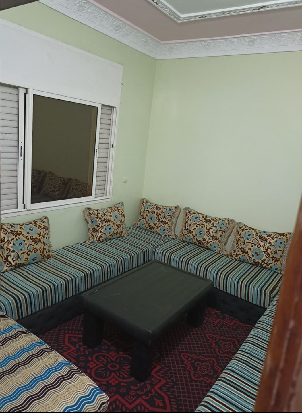 A cozy seating area features a large, low table at its center surrounded by a U-shaped couch. The couch is upholstered in a striped fabric with colorful cushions. Natural light enters through a window dressed with shutters, creating a relaxed ambiance.