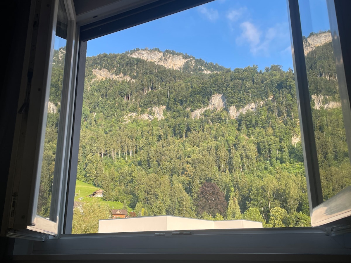 An open window provides a view of lush green mountains, framed by surrounding trees. The sky above is clear, with soft clouds enhancing the serene setting.