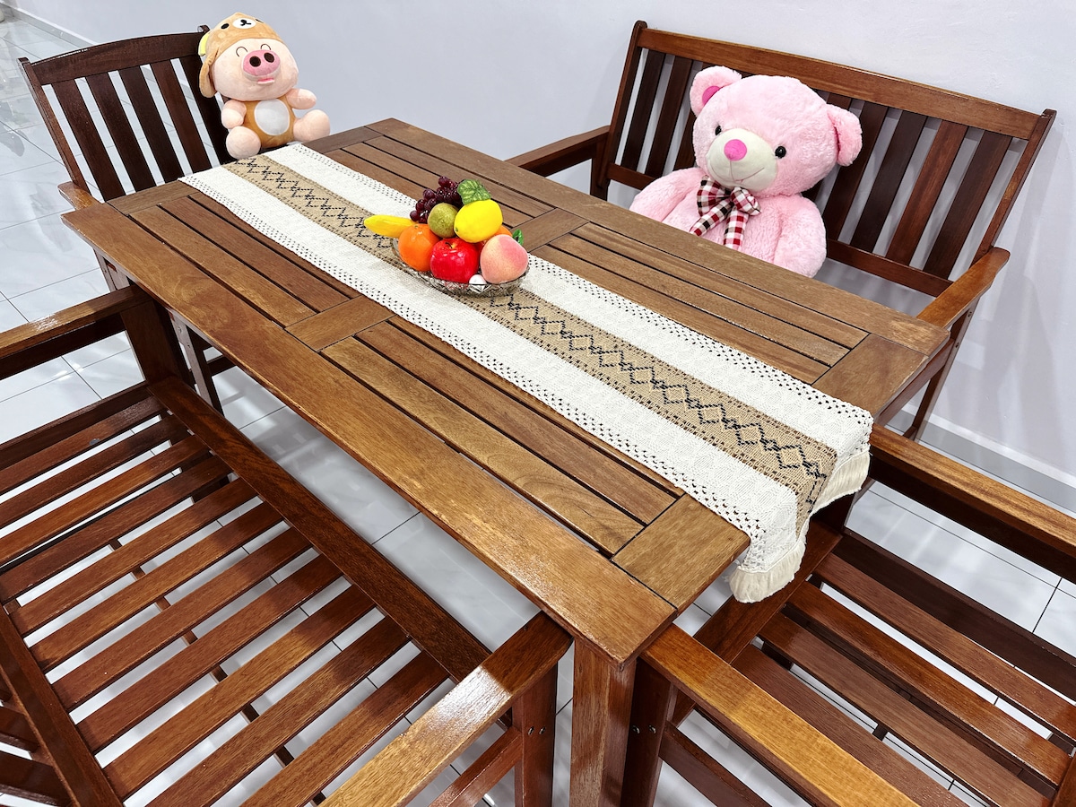 A wooden dining table is set with a decorative runner and a variety of fresh fruits arranged in the center. Two cozy stuffed animals sit on opposite chairs, adding a playful touch to the setting. The surrounding area is bright and uncluttered.