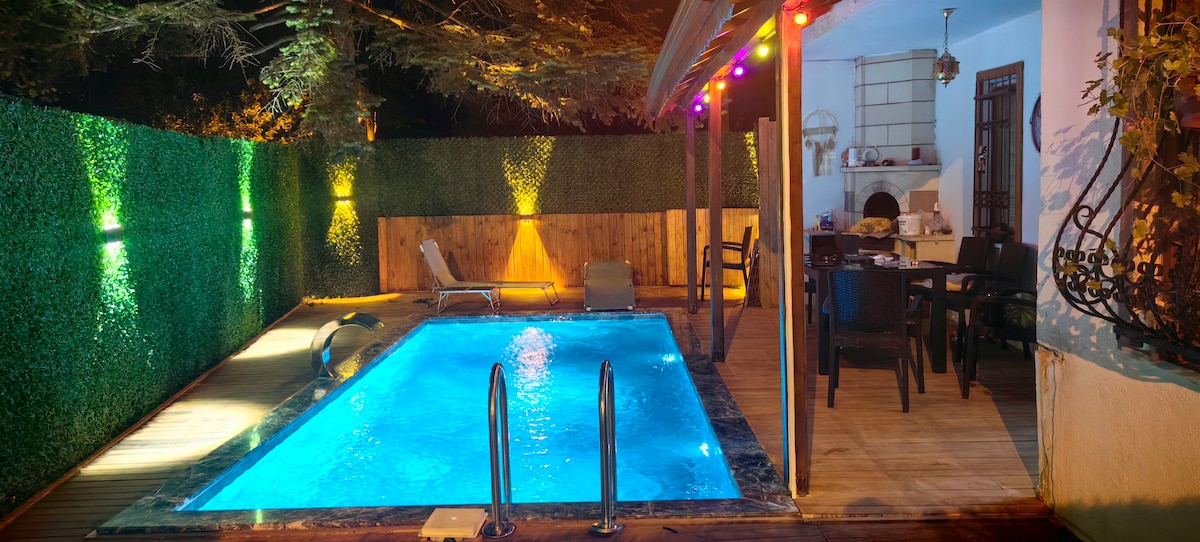A private outdoor space features a shimmering pool surrounded by lush green foliage and softly glowing lights. Comfortable seating arrangements and a dining area are visible under a covered patio, creating an inviting environment for relaxation and social gatherings.