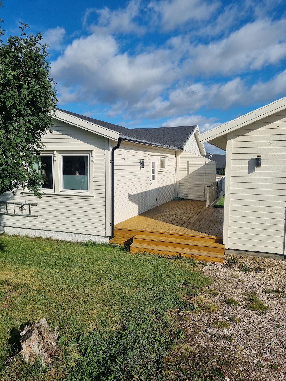 A cozy entrance is featured, leading to a wooden deck that connects two light-colored structures. The exterior is complemented by green grass and a few scattered stones. The sky above reflects a mix of clouds, contributing to a serene outdoor setting.