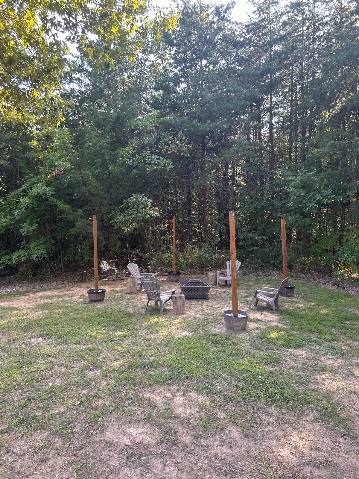 A fire pit area is surrounded by several seating options, including wooden and metal chairs. The clearing features a grassy surface, bordered by tall trees providing privacy. Four wooden posts are positioned around the fire pit, enhancing the rustic charm of the outdoor space.