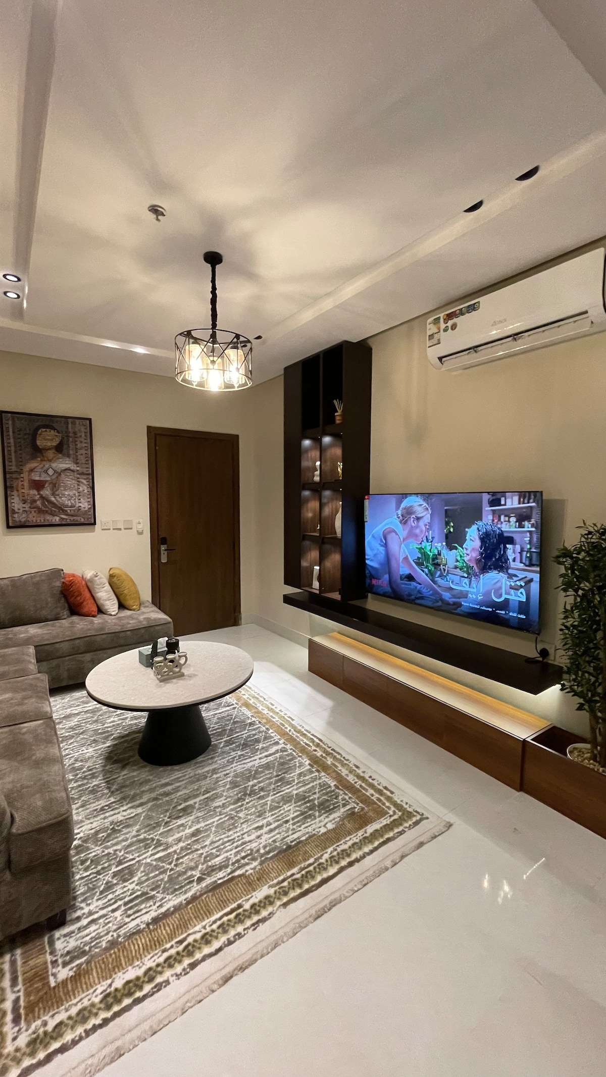 A modern living area showcases a comfortable sectional sofa adorned with colorful cushions. A round coffee table is centered on a patterned area rug, while a wall-mounted television is framed by minimalist cabinetry. Soft lighting is provided by a stylish ceiling fixture.