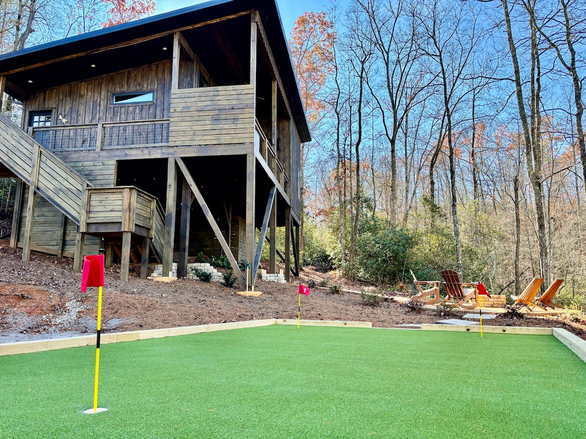 A private putting green is lined with red flags and framed by lush green turf. In the background, a treehouse structure stands elevated on stilts, surrounded by wooded scenery. Cozy seating areas are visible nearby, creating an inviting outdoor space.