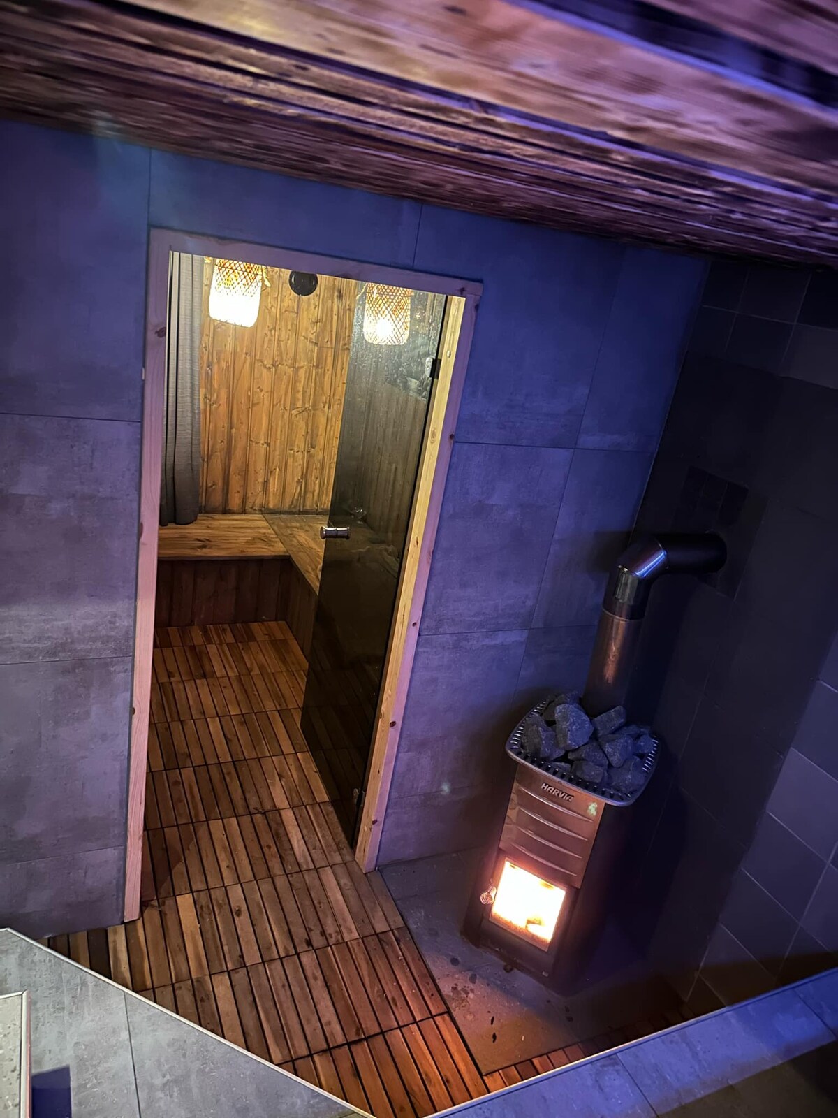 The entrance to a wooden sauna is visible, featuring warm wooden benches and soothing lighting. A stone stove sits beside the entrance, with logs stacked nearby. The floor is covered in wooden slats, creating a natural aesthetic and inviting environment for relaxation.