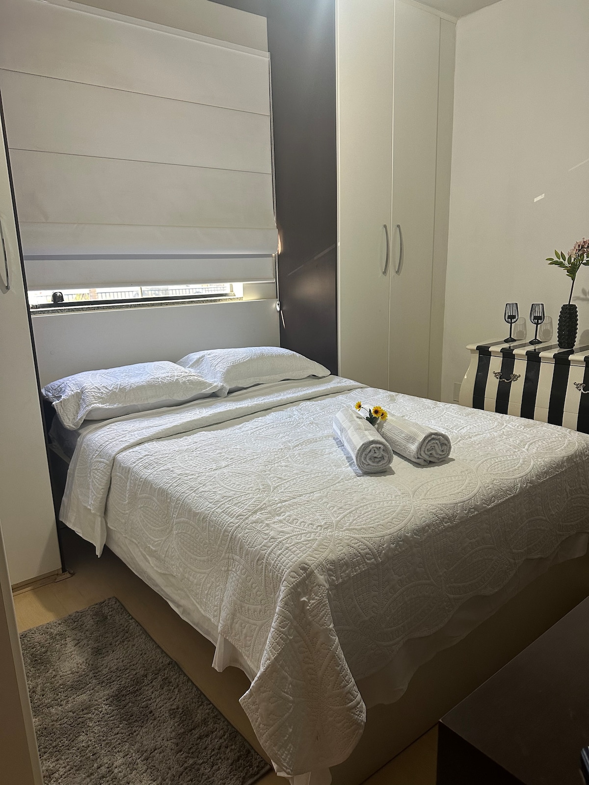 A cozy sleeping area features a spacious bed covered with a white textured blanket. Neatly arranged towels and a sunflower are placed at the foot of the bed. Soft natural light filters through a window dressed with a light fabric shade, illuminating the room's minimalistic decor.