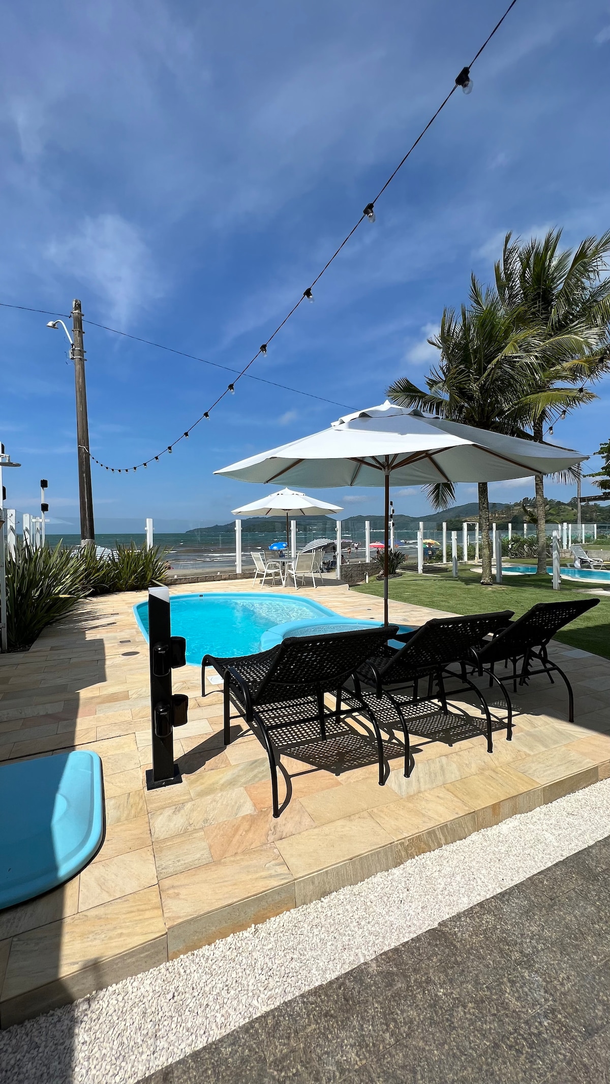 A swimming pool is framed by a stone patio, with black lounge chairs positioned nearby under large white umbrellas. Green palm trees and additional outdoor seating can be seen in the background, while string lights are suspended overhead against a clear blue sky.