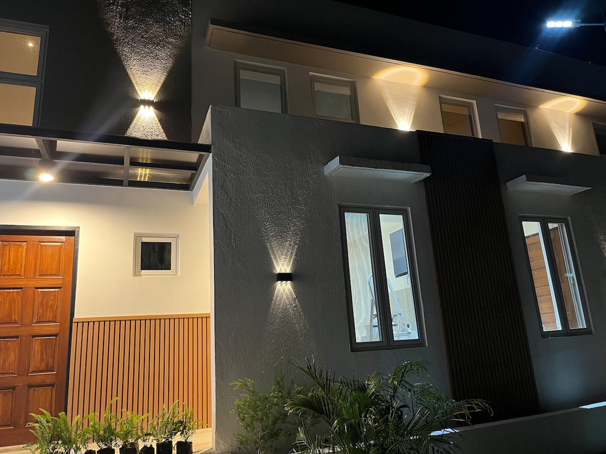 A modern two-story home is illuminated at night, highlighting its clean lines and architectural features. Large windows reflect soft light, while a wooden front door adds a natural element. Subtle landscaping with small plants complements the entrance.