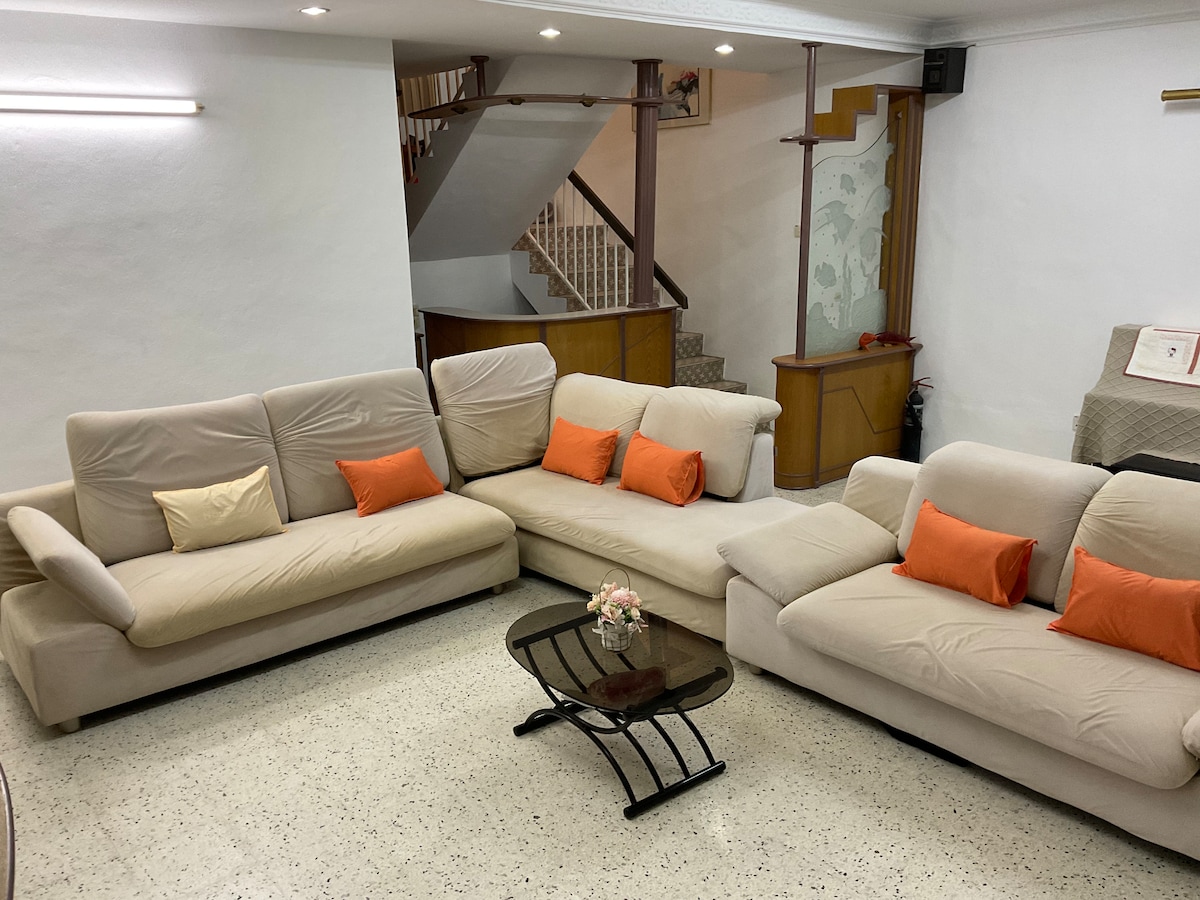 A spacious living room is showcased with a large sectional sofa, featuring soft beige upholstery and accent cushions in shades of orange. A sleek glass coffee table rests in front, accompanied by a small floral arrangement. A staircase leads to the upper level in the background.