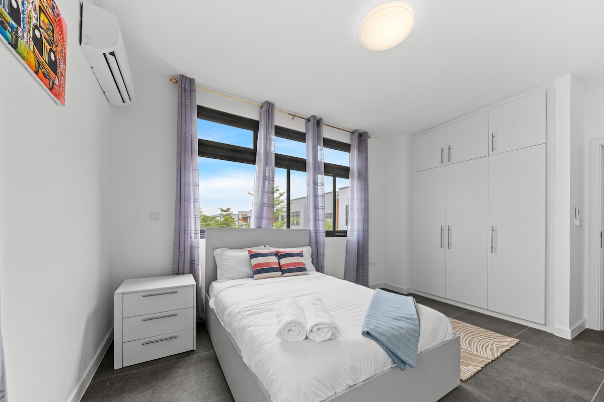 غرفة نوم ثالثة مشرقة بسرير مزدوج ومكيف هواء وبياضات نظيفة. توفر النوافذ الكبيرة إضاءة طبيعية وستائر خصوصية. خزانة ملابس واسعة وصندوق بجانب السرير للراحة. تشطيبات نظيفة وحديثة لإقامة مريحة.